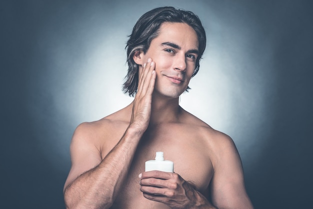 Sentirse fresco y limpio. Retrato de hombre sin camisa joven guapo mirando a cámara y aplicando loción para después del afeitado en la cara mientras está de pie contra el fondo gris