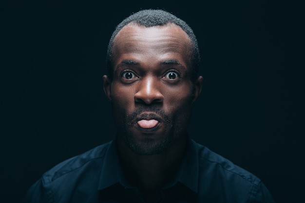 Sentirse juguetón. Retrato de alegre joven africano sacando la lengua y mirando a la cámara con una sonrisa mientras está delante de un fondo negro