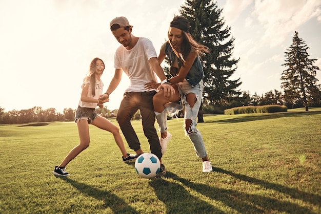 Sentirse libre con amigos. Longitud total de jóvenes sonrientes en ropa casual corriendo mientras juegan al fútbol al aire libre