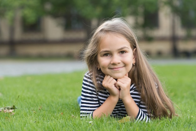 Sentirse libre. Sonrisa de niño feliz tumbado en la hierba verde. Mirada de belleza. Look de moda de niña pequeña. Peluquería. Cuidado de la piel y del cabello. Ropa de ninos. Vacaciones de verano.