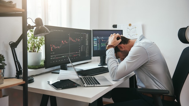 Sentirse triste y deprimido joven empresario o comerciante en ropa formal manteniendo la cabeza entre las manos mientras trabaja