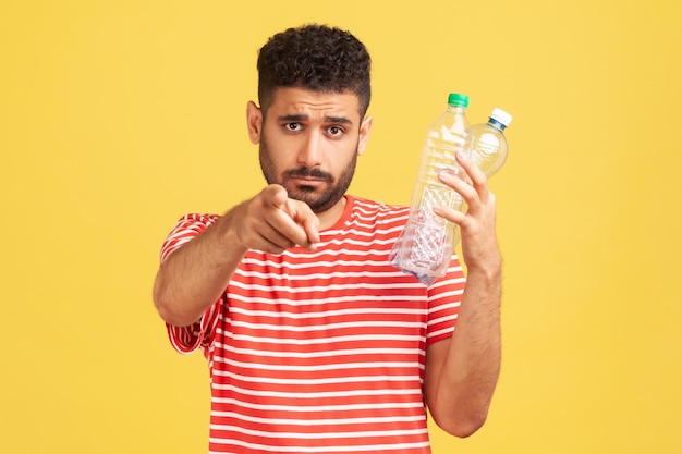 Separe seu lixo. Homem barbudo sério em camiseta listrada segurando garrafa de plástico e apontando o dedo para a câmera, se preocupando com o meio ambiente. Tiro de estúdio interior isolado em fundo amarelo