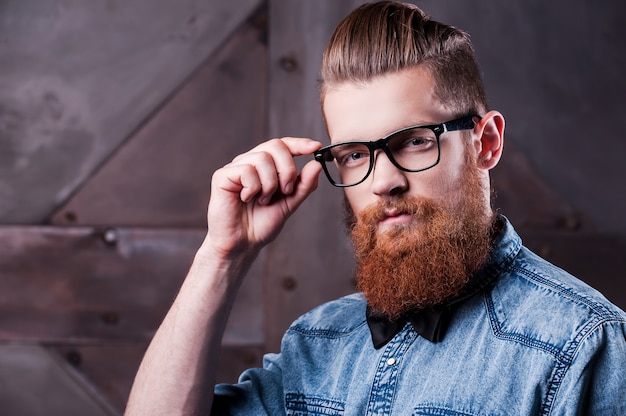 Ser retro es tendencia. Hombre barbudo joven hermoso que lleva a cabo la mano en los vidrios y que mira la cámara