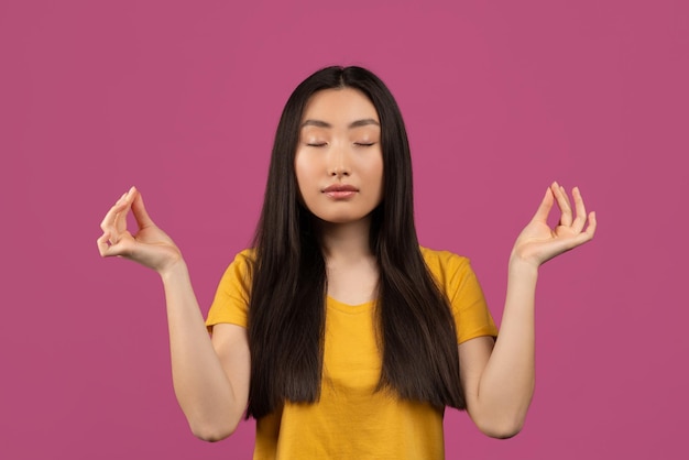 Serena jovem asiática com olhos fechados meditando desfrutando de silêncio e relaxamento sobre violeta