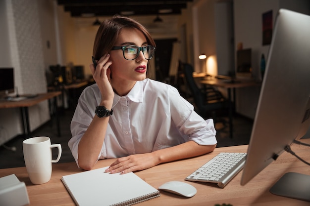 Séria atraente jovem empresária em óculos escrevendo e usando o computador no escritório