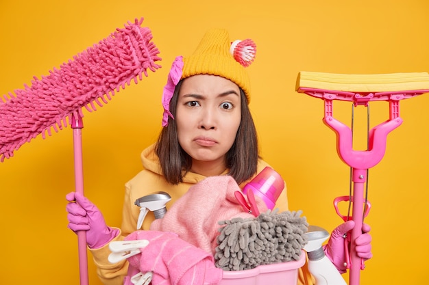 Séria descontente jovem asiática faz limpeza de primavera olha escrupulosamente para a câmera usa chapéu, luvas de borracha com capuz e detém dois esfregões em poses de mãos com cesto de roupa suja e detergentes internos.