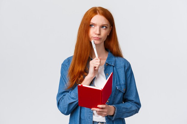 Séria e determinada mandona ruiva bonitinha com caderno, segurando a caneta, sorria pensativa e desviou o olhar ao pensar, lembre-se do que comprar, prepare mantimentos ou lista de tarefas,