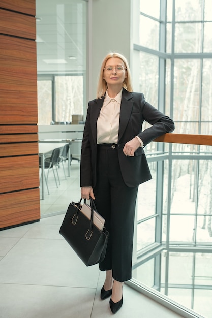 Séria elegante mulher de negócios madura em um terno preto em pé na grade e segurando uma bolsa enquanto olha para a câmera