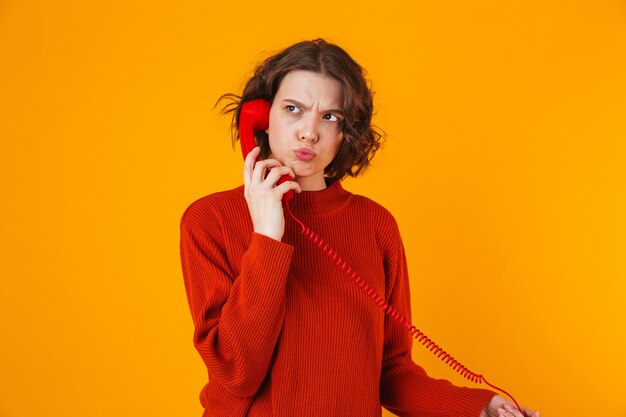 séria jovem bonita posando isolada na parede amarela, falando por telefone.