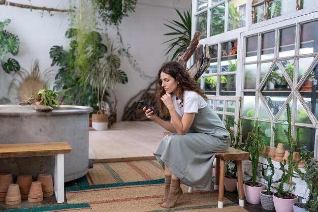 Foto seria joven jardinera mujer propietaria sosteniendo un teléfono inteligente leyendo noticias de los medios sentada en el jardín de casa