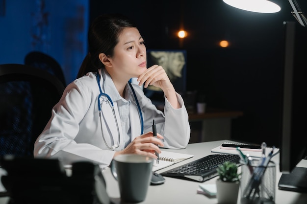 Una seria y reflexiva doctora china asiática que trabaja con una computadora en la mesa en un lugar de trabajo oscuro. el personal médico de la dama pensativa frunciendo el ceño y mirando la pantalla de la pc pensando en la solución del problema del paciente.