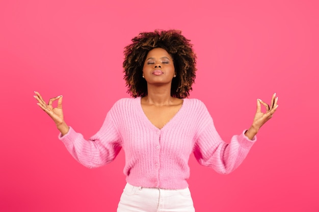 Seria y tranquila joven mujer negra rizada en meditación informal con los ojos cerrados aislada en un fondo rosa