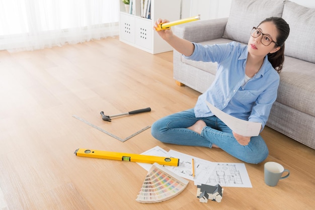 seriamente bonita dona de casa sentada na sala de estar piso de madeira segurando engenheiro fita régua ferramenta medição casa interior renovar design com foto de vista de alto ângulo.