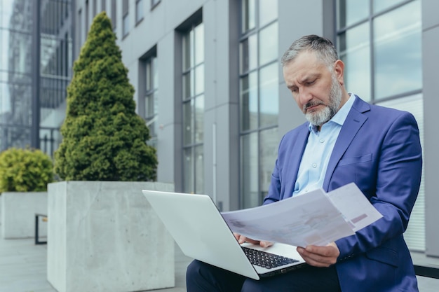 Serio, apuesto y exitoso hombre de negocios masculino trabaja con una computadora portátil y los documentos concluyen en línea