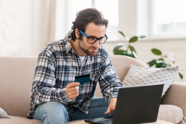 Serio concentrado joven caucásico adicto a las compras en gafas se sienta en el sofá con computadora y tarjeta de crédito