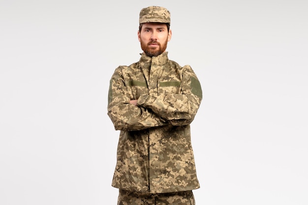 Serio hombre barbudo soldado vestido con uniforme militar con los brazos cruzados aislado en el fondo