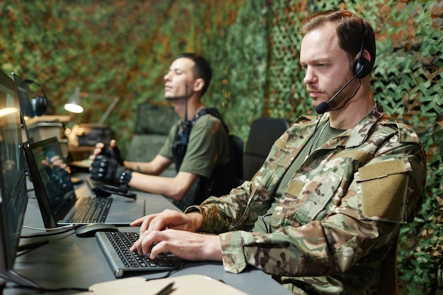 Foto serio joven operador masculino en uniforme de camuflaje escribiendo en el teclado