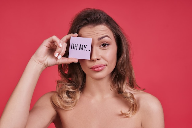 ¿En serio? Mujer joven aburrida que cubre un ojo con un cartel pequeño y mirando a la cámara mientras está de pie contra el fondo rosa