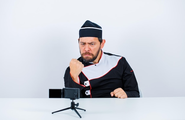 Sério, o chef blogueiro está levantando o punho sentado na frente de sua mini câmera no fundo branco