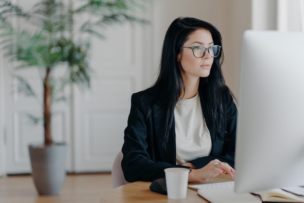 Seriöse, elegante Geschäftsfrau im Coworking Space konzentriert sich auf den Monitor, trinkt Kaffee und analysiert Daten