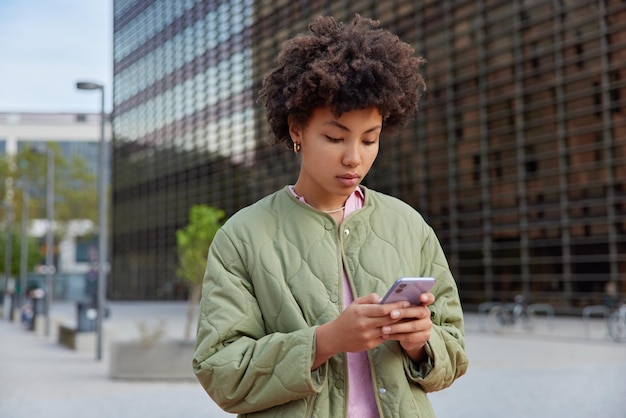 Foto seriöse lockige frau in freizeitjacke sendet textnachrichten über smartphone nutzt drahtloses internet zum teilen von blogging oder teilen von veröffentlichungsspaziergängen im freien im städtischen umfeld moderne technologien