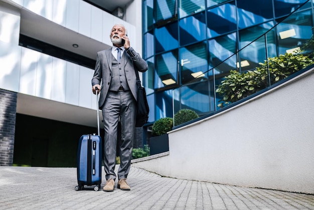 Seriöser älterer Geschäftsmann mit Koffer, der mit dem Smartphone gegen ein Gebäude spricht