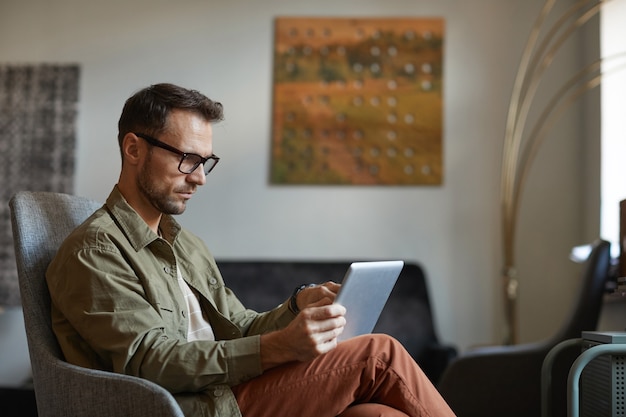 Seriöser Designer in Brillen, die auf Sofa sitzen und online auf digitalem Tablett im modernen Büro arbeiten