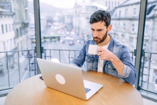 Seriöser Geschäftsmann, erster Gast in einem Café, der in einem Café sitzt und auf den Computerbildschirm schaut und Online-Nachrichten liest, die heute beginnen, morgens heißen Kaffee zu trinken