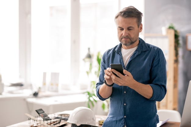 Seriöser Ingenieur in Freizeitkleidung, der während der Arbeit im Büro durch Kontakte in seinem Smartphone blättert