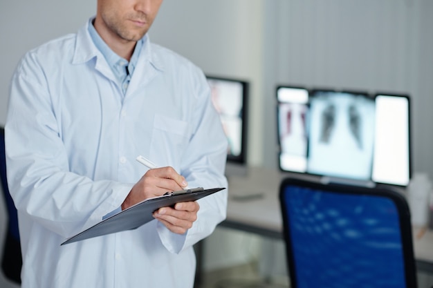 Seriöser Lungenarzt, der die Diagnose des Patienten aufschreibt, nachdem er seine Lungenröntgenaufnahme auf dem Computerbildschirm überprüft hat