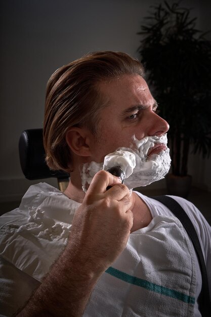 Seriöser männlicher Kunde in weißer Kleidung trägt weißen Rasierschaum auf den Bart auf, während er während eines Termins in einem hellen, modernen Friseursalon sitzt
