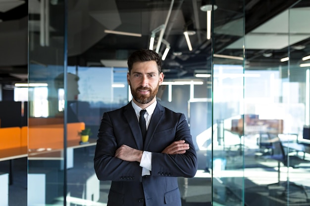 Seriöser und selbstbewusster Geschäftsmann mit verschränkten Armen, der in einem modernen Büro in die Kamera blickt