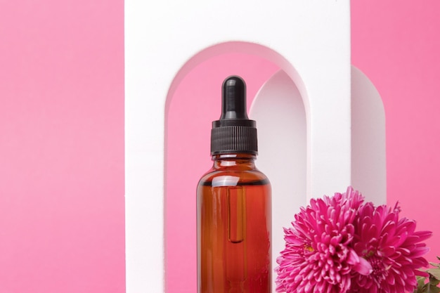 Serum mit Blumenextrakten für die Hautpflege im Bogen Naturkosmetik in Glasflasche mit Pipette und rosa Blüten auf rosa Hintergrund Spa-Konzept für Gesichts- und Körperpflege