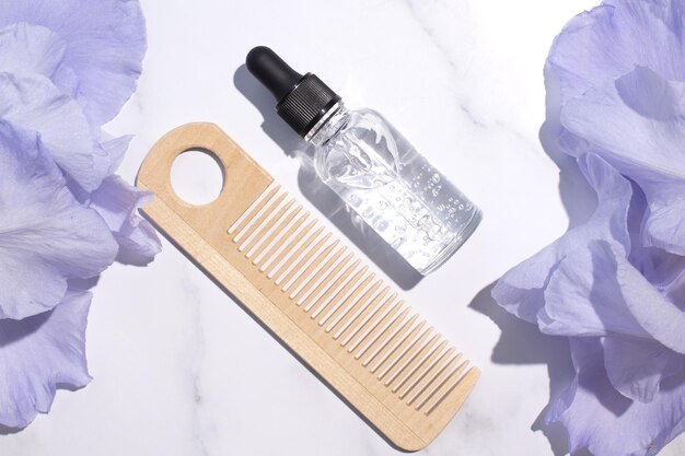 Serum mit Blumenextrakten für Hautpflege und Haarkamm aus Holz Naturkosmetik in Glasflasche mit Pipette und Irisblüten auf blauem Hintergrund Spa-Konzept für Gesichts- und Körperpflege