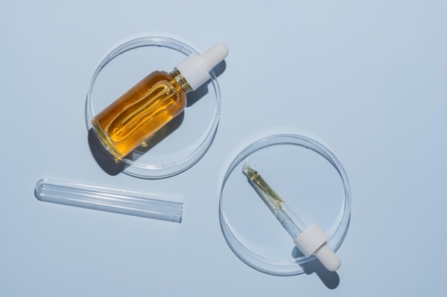 Serumflasche und kosmetische Pipette mit Öl in Petri-Schüssel auf blauem Hintergrund Stilvolles Konzept von organischen Essenzen Schönheits- und Gesundheitsprodukten Moderne Apotheke Close Up