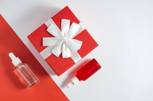 Serumglassflaschen mit Pipette und roter Geschenkbox auf rot-weißem Hintergrund mit Kopierraum moderne Zusammensetzung Minimalistische Haut- und Körperpflege Winterverkaufskonzept Flatlay Knolling