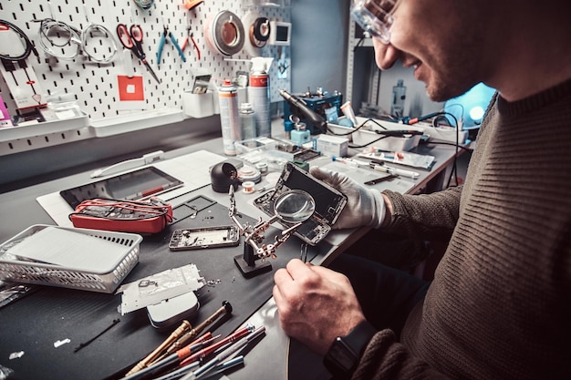 Serviceman usa lupa y pinzas para reparar el teléfono inteligente dañado en el taller electrónico.