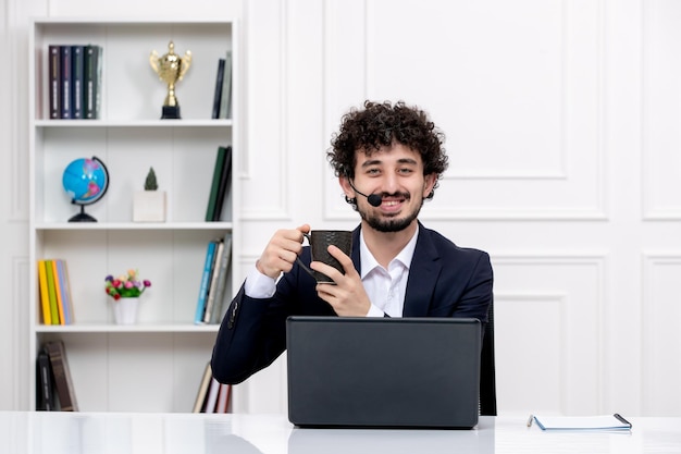 Servicio al cliente apuesto hombre rizado en traje de oficina con computadora y auriculares sonriendo con café