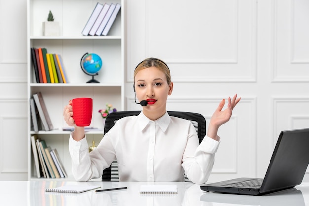 Servicio al cliente linda chica rubia con camisa blanca con computadora portátil y auriculares sosteniendo una taza de café