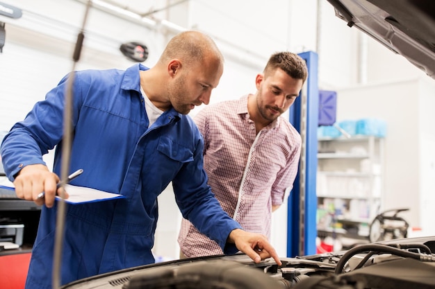 servicio automático, reparación, mantenimiento y concepto de personas - mecánico con portapapeles y hombre o propietario mirando el motor de camión roto en la tienda