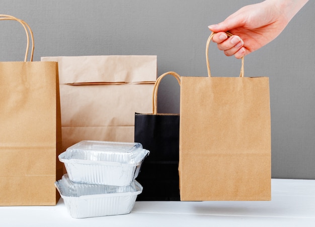 Servicio de comida a domicilio. Paquete de bolsa de papel artesanal marrón en mano femenina. Embalaje de entrega. Envases de aluminio y paquetes de papel sobre fondo gris de mesa. Comida para llevar.