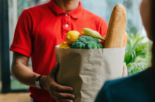 Servicio de entrega de alimentos guapo hombre en camisa roja con bolsa de set de alimentos frescos al cliente en la puerta de su casa, entrega urgente