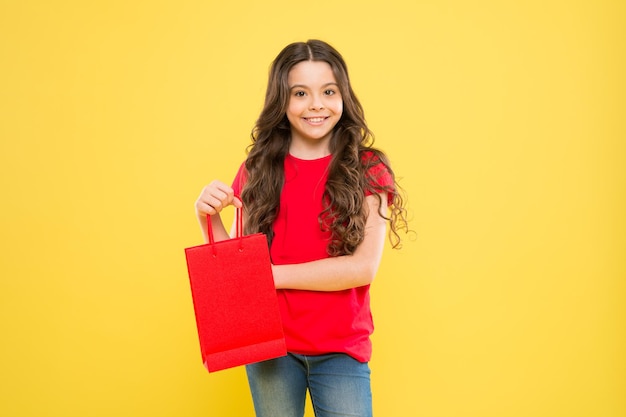 Servicio de entrega niño sobre fondo amarillo niño de compras con bolsa de compras niña feliz con cabello largo y rizado pequeña belleza adicta a las compras Regalo de vacaciones niña comprando en línea comprador feliz