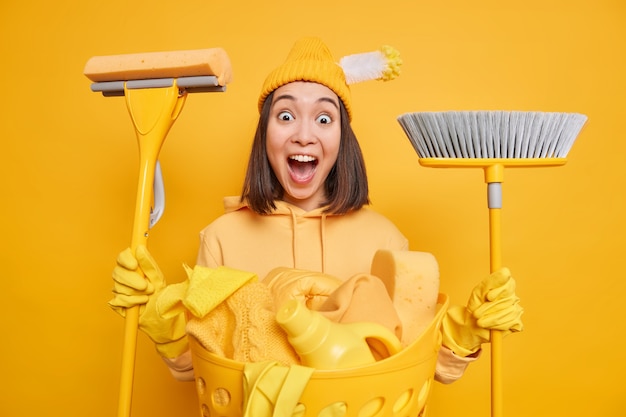 Servicio de limpieza concepto de tareas domésticas y limpieza. La mujer asiática positiva sostiene la fregona y la escoba se preocupa por la nueva casa hace las tareas del hogar vestidas casualmente aisladas sobre fondo amarillo del estudio