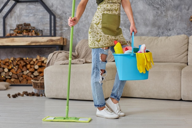 Servicio de limpieza. Cubo con esponjas, botellas de productos químicos y bastoncillo para trapear. Guantes de goma y toalla. Equipo para el hogar. Imagen recortada de una mujer con una fregona en la sala de estar