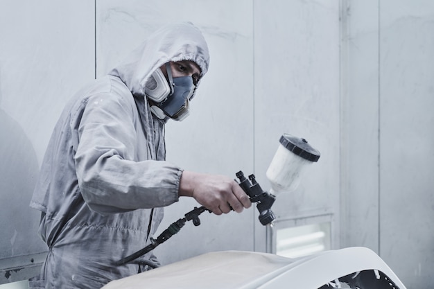 Foto servicio de pintura y reparación de automóviles. mecánico de automóviles en mono blanco pinta coche con pulverizador de aerógrafo en la cámara de pintura