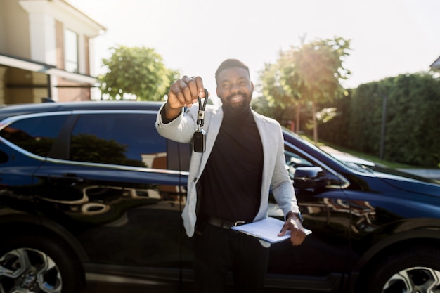 Serviço de aluguel de carros. Vendedor de homem africano feliz ou cliente segurando a chave e sorrindo perto de carro preto novo. Foco na tecla