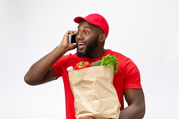 Serviço de entrega - retrato de um homem afro-americano bonito entregador ou mensageiro com um pacote de mantimentos e falando no celular para verificar o pedido.