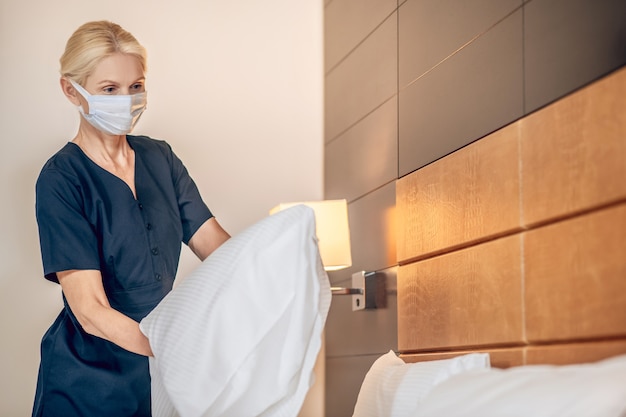 Serviço de quarto. Mulher loira com máscara protetora fazendo cama em um quarto de hotel