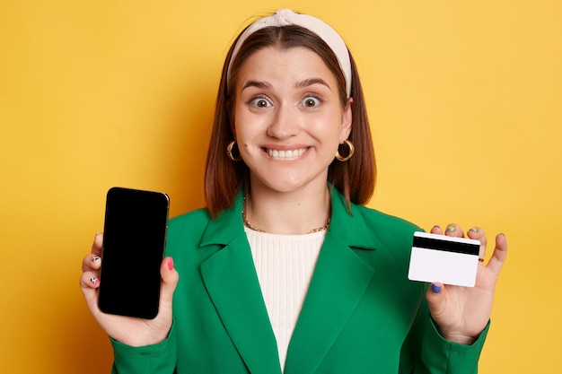 Serviços bancários por telefone Pagamentos on-line Mulher feliz encantada vestindo jaqueta verde posando isolada sobre fundo amarelo mostrando telefone celular com tela vazia e cartão de crédito